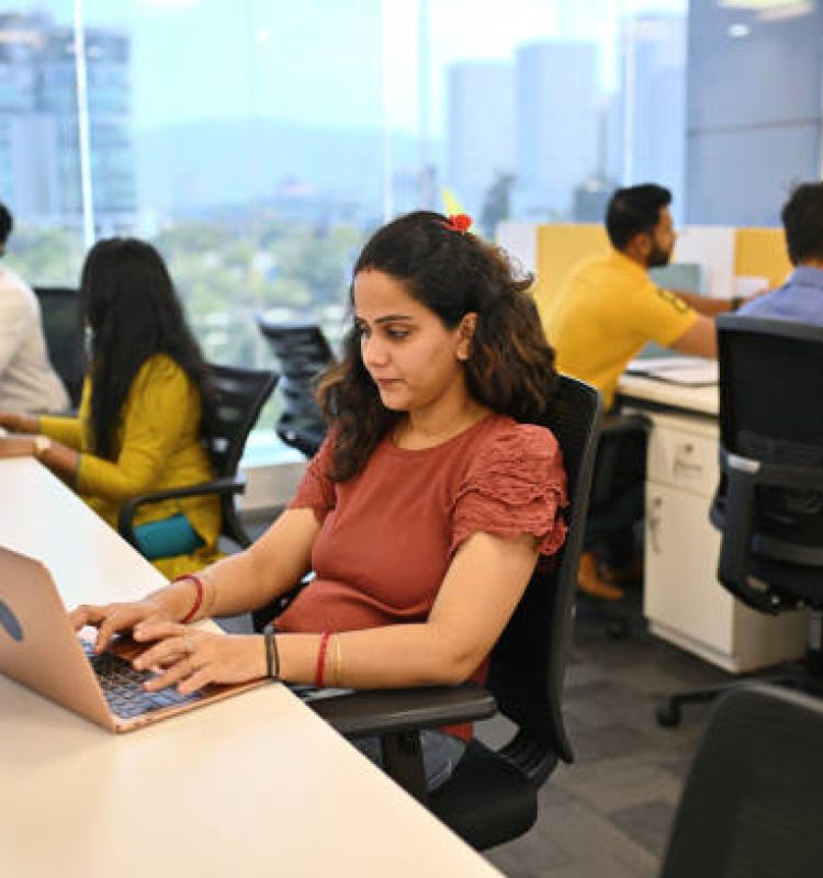 Photo portrays a typical office setup with a female employee working on a laptop while few are discussing and others are working sitting on their desks in an office cubicle. The photo conveys a sense of productivity in the workplace.
